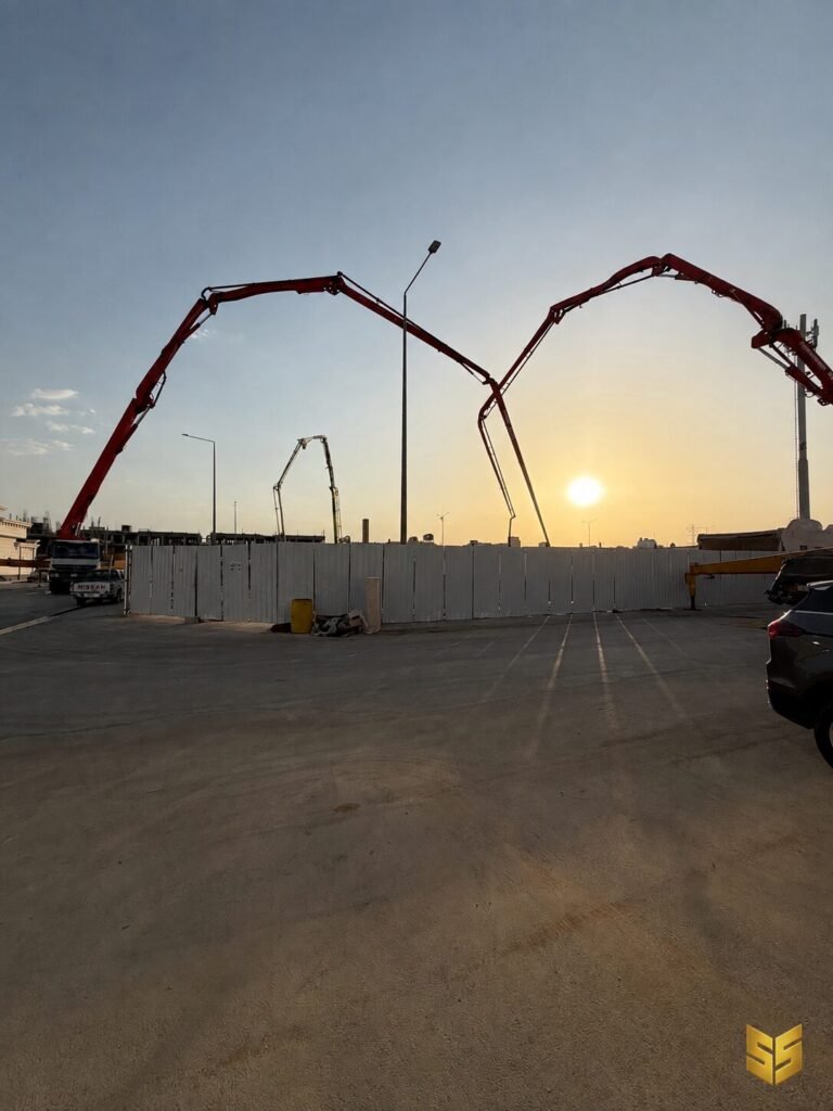 أذرع مضخات خرسانة ممتدة فوق موقع مشروع عند غروب الشمس استعدادًا لصبة اللبشة في الرياض